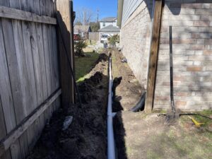 Trench for drainage installation alongside a house, showcasing preparation for landscaping in Houston's soil and climate.