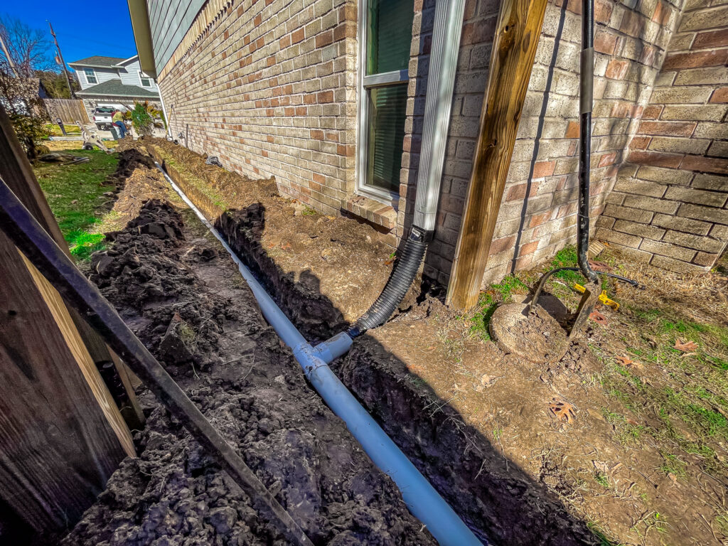 Irrigation system installation in progress alongside a brick home in Tomball, showcasing a trench with piping and tools, highlighting Danny's Landscaping's commitment to tailored outdoor solutions.
